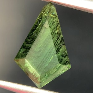 May include: A faceted, kite-shaped gemstone with a vibrant green hue. The gem is held by tweezers, showcasing its geometric cut and internal patterns. The gemstone's facets reflect light, highlighting its clarity and color.