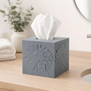 May include: A gray, cube-shaped tissue box with a large, embossed floral design on two sides. White tissues are pulled from the top opening. The box is on a wooden surface, with a blurred background.