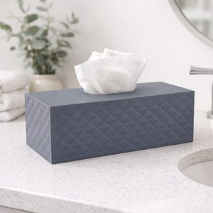 May include: A rectangular, slate-grey tissue box with a geometric diamond pattern. White tissues protrude from the top. The box is on a white countertop in a bathroom, with a sink and mirror in the background.