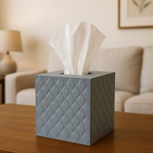 Puede incluir: Un dispensador de pañuelos de papel gris geométrico con un patrón de diamantes. Los pañuelos blancos sobresalen de la abertura superior. La caja es cuadrada y está sobre una superficie de madera. El fondo incluye una lámpara, un sofá y una imagen enmarcada.