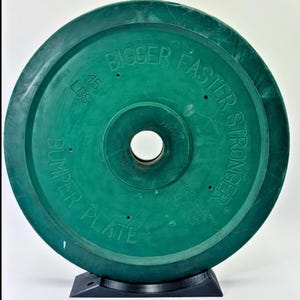 以下が含まれることがあります： 重量挙げ用の緑色のバンパープレート。表面には「Bigger, Faster, Stronger」と「45 LBS」の文字がエンボス加工されています。プレートには中央に穴があり、黒いスタンドで支えられています。「Bumper Plate」の文字も確認できます。