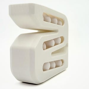 May include: A cream-colored, geometric object with rounded edges and multiple horizontal openings. Each opening contains three white spheres. The design is modern and abstract, with a clean, minimalist aesthetic.