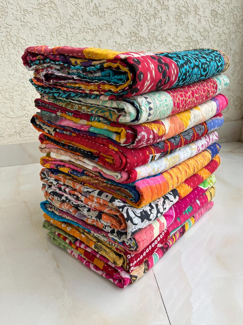 May include: A pile of vibrant patchwork quilts. The quilts display a range of patterns and colours, including red, blue, yellow, and pink. The quilts are neatly folded, highlighting the detailed stitching and design.