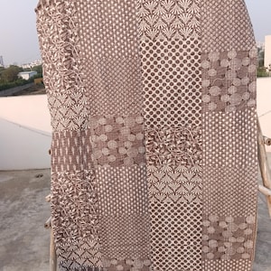 May include: A brown and white patchwork quilt featuring diverse patterns such as polka dots, floral motifs, and geometric shapes. The quilt is displayed outdoors, draped over a wooden structure, highlighting its detailed design and texture.