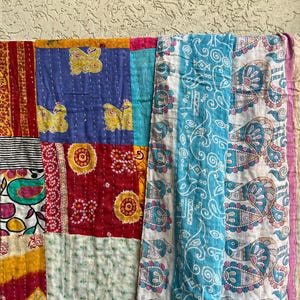 May include: A vibrant patchwork quilt with a mix of patterns and colours. The quilt displays squares of red, blue, yellow, and turquoise fabric. Some squares have butterfly and floral designs. The quilt is hanging on a blue rod.