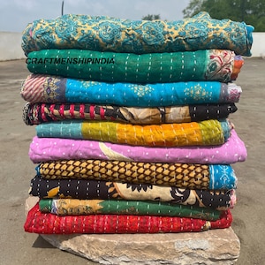 Op de afbeelding: Een stapel kleurrijke, bedrukte quilts. De quilts tonen verschillende ontwerpen, waaronder paisley- en bloempatronen, in tinten van teal, groen, roze, geel en rood. De tekst "CRAFTSMANSHIPINDIA" is zichtbaar.