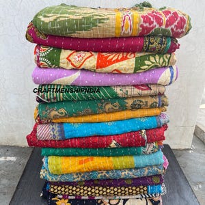 Puede incluir: Una pila de colchas coloridas y estampadas. Las colchas presentan una variedad de colores, incluyendo verde, rojo, amarillo, azul y morado. Las colchas están hechas de un material suave y están apiladas. El texto "CRAFTSHIPINDIA" es visible.