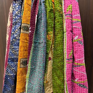 Op de afbeelding: Een collectie van meerdere lange, rechthoekige stoffen sjaals met diverse patronen en kleuren, waaronder diepblauw, mosterdgeel, bordeauxrood, groenblauw, groen en felroze, allemaal voorzien van zichtbaar wit rijgstiksel.