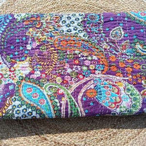Op de afbeelding: Een gevouwen, gewatteerde textiel met een levendig paisley patroon. De quilt heeft een rijke paarse basiskleur met accenten van oranje, blauwgroen, groen en wit. Het ontwerp bevat bloem- en druppelvormen en is gestikt met witte draad.