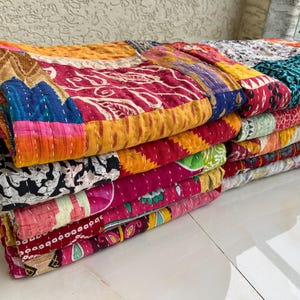 May include: A stack of colourful, patchwork quilts. The quilts feature a variety of patterns and colours, including red, yellow, blue, and pink. Each quilt is made of multiple fabric squares stitched together with white thread. The quilts are stacked on top of each other.