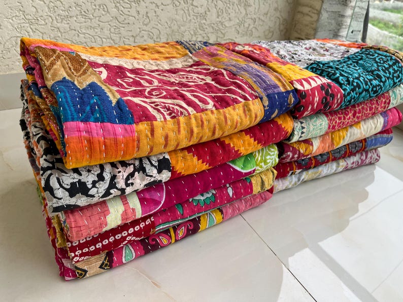 May include: A stack of colourful, patchwork quilts. The quilts feature a variety of patterns and colours, including red, yellow, blue, and pink. Each quilt is made of multiple fabric squares stitched together with white thread. The quilts are stacked on top of each other.