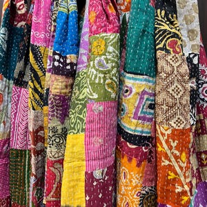 May include: A multi-coloured patchwork scarf featuring various fabric squares with diverse patterns including floral, geometric, and paisley prints in shades of pink, yellow, green, blue, and orange, all joined together with visible white running stitch.