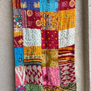 May include: A patchwork textile with a variety of colourful squares. The squares are in shades of red, blue, yellow, and green, with various patterns and textures. The textile is hanging on a blue rod.