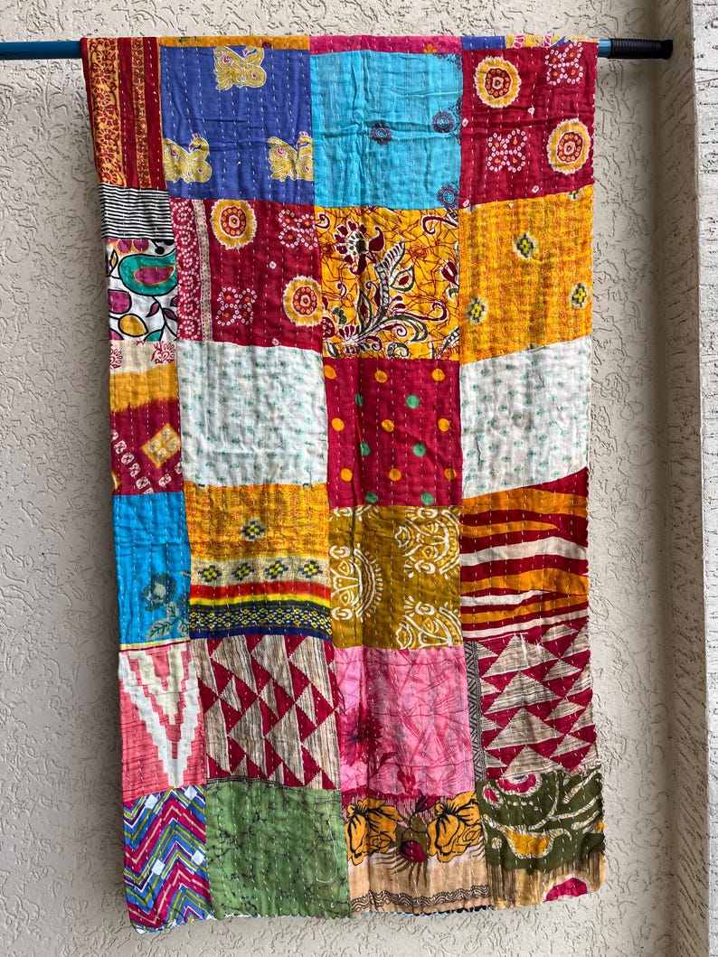 May include: A patchwork textile with a variety of colourful squares. The squares are in shades of red, blue, yellow, and green, with various patterns and textures. The textile is hanging on a blue rod.