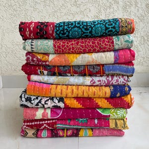 May include: A pile of vibrant patchwork quilts. The quilts are made from a variety of fabrics in red, blue, yellow, and pink. Each quilt is folded and stacked, displaying diverse patterns and textures. The quilts are likely hand-stitched.