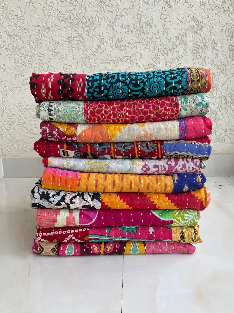 May include: A pile of vibrant patchwork quilts. The quilts are made from a variety of fabrics in red, blue, yellow, and pink. Each quilt is folded and stacked, displaying diverse patterns and textures. The quilts are likely hand-stitched.