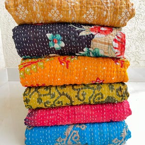 May include: A stack of colorful, rolled kantha quilts. The quilts are in shades of yellow, black, orange, green, pink, and blue. Each quilt features a unique floral or patterned design and is stitched with white thread.