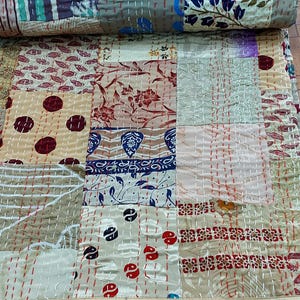 Puede incluir: Un edredón de patchwork con cuadrados de tela estampada en tonos beige, marrón, azul y rojo. El edredón está cosido a mano con hilo blanco, y algunas secciones están enrolladas.