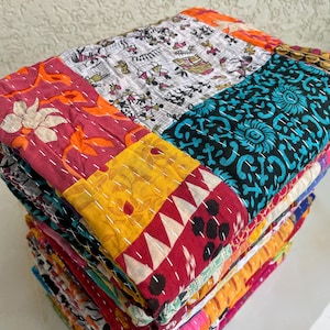 May include: A stack of vibrant patchwork quilts. Each quilt displays a unique mix of patterns and colours, including floral, geometric, and abstract designs. The quilts are made from various fabrics and stitched together with white thread.