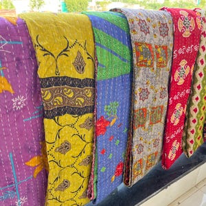 May include: A selection of patterned textiles, possibly quilts or throws, are displayed. The fabrics feature a variety of designs, including floral and geometric patterns, in shades of purple, yellow, blue, red, and green. The textiles are folded, showcasing the diverse patterns.