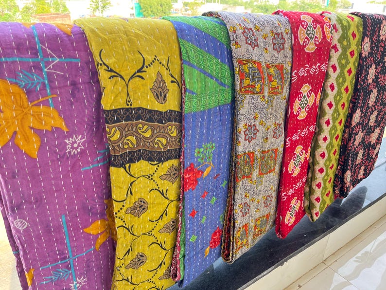 Pu&ograve; includere: Una collezione di tessuti colorati e fantasia, probabilmente trapunte o coperte, &egrave; esposta. I tessuti presentano una variet&agrave; di disegni, tra cui motivi floreali e geometrici, nelle tonalit&agrave; del viola, giallo, blu, rosso e verde. I tessuti sono piegati e disposti.