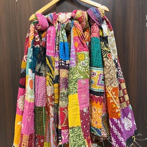 Puede incluir: Una colcha de patchwork que presenta una vibrante variedad de cuadrados de tela multicolores con diversos patrones florales, geométricos y de cachemira. El textil está construido con costuras blancas visibles y cuelga de una percha de madera.