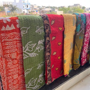 Könnte beinhalten: Eine Sammlung farbenfroher, gemusterter Textilien, wahrscheinlich Decken oder Überwürfe, wird im Freien gezeigt. Die Textilien weisen eine Vielzahl von Designs auf, darunter geometrische Muster und florale Motive, in Rot-, Grün-, Gelb- und Rosatönen.