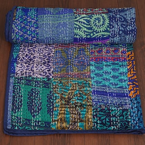 Könnte beinhalten: Eine Patchwork-Steppdecke in Blau-, Türkis- und Grüntönen mit verschiedenen gemusterten Stoffquadraten. Die Steppdecke ist mit weißem Faden handgenäht und hat einen aufgerollten Abschnitt oben. Die Kanten sind mit einem einfarbigen blauen Rand versehen.