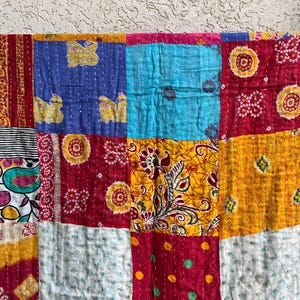 May include: A colourful patchwork quilt made from squares of fabric. The quilt has blue, red, yellow, and white squares with floral and butterfly patterns. It is hanging on a blue rod.