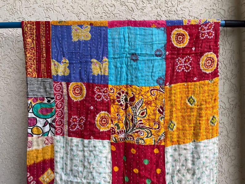 May include: A colourful patchwork quilt made from squares of fabric. The quilt has blue, red, yellow, and white squares with floral and butterfly patterns. It is hanging on a blue rod.