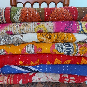 Puede incluir: Una pila de edredones coloridos y estampados. Los edredones presentan varios diseños, incluidos patrones florales y geométricos, en tonos de rojo, rosa, amarillo, verde y azul. Cada edredón está doblado y apilado uno encima del otro.