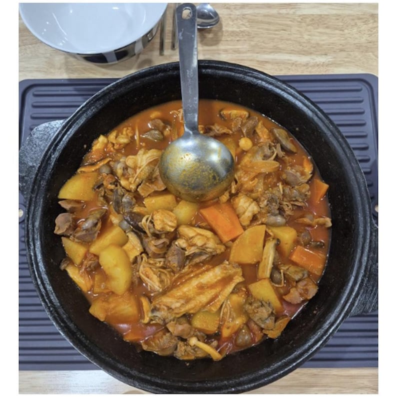 May include: A close-up shot of a hearty stew in a black pot. The stew contains chicken pieces, potatoes, carrots, and mushrooms in a rich, reddish-brown broth. A metal ladle rests in the stew. A white bowl is in the background.