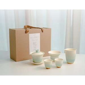 Op de afbeelding: Een theeset bestaande uit een gaiwan met deksel en schoteltje, een theepot en twee kleine kopjes, allemaal met een wit naar lichtblauw kleurverloop. De set wordt gepresenteerd in een bruine kartonnen doos met het label Jeju Beach Tea Set.