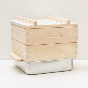 May include: A wooden steamer with two stacked, light-coloured wooden boxes and a stainless steel base. The boxes have recessed handles and are topped with two wooden bars. The steamer is designed for cooking.