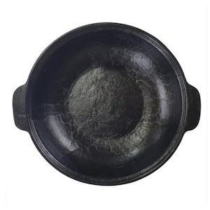 May include: A black cast iron cooking pot with a textured interior and two small handles on either side. The pot has a rounded shape and is designed for cooking. The pot is set against a white background.