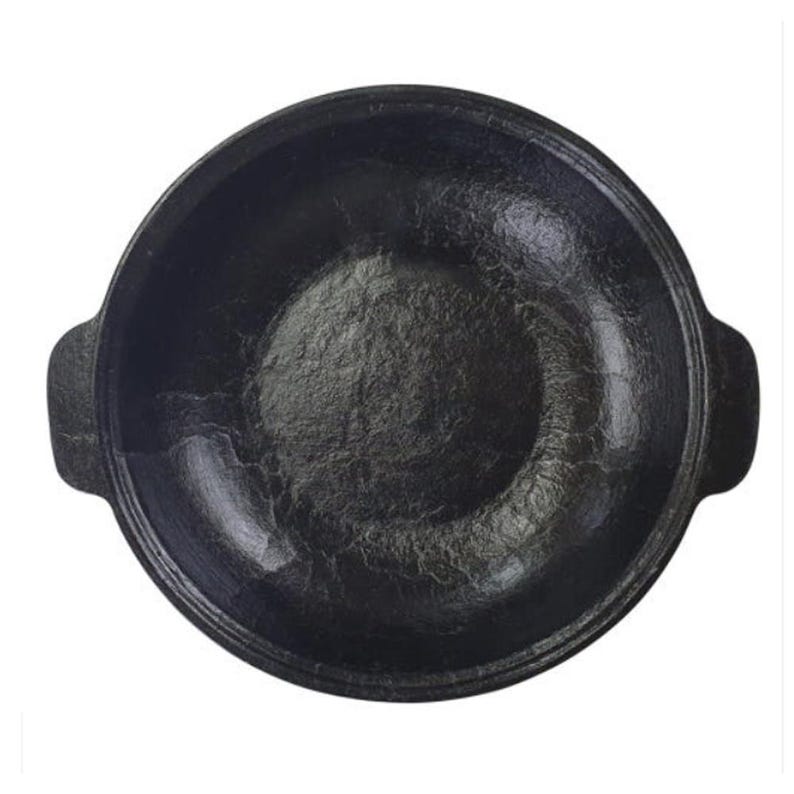 May include: A black cast iron cooking pot with a textured interior and two small handles on either side. The pot has a rounded shape and is designed for cooking. The pot is set against a white background.