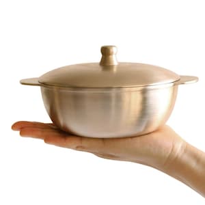 May include: A small, round, metallic bowl with a brushed gold finish, featuring a matching lid with a central knob handle and two small side handles. The bowl has a smooth, curved body and a polished, reflective surface throughout its exterior.
