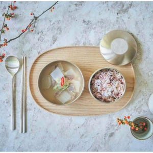 May include: A traditional Korean meal is presented on a wooden tray. The arrangement includes a bowl of soup with vegetables, a bowl of rice, and a covered dish. Silverware and a small decorative vase with flowers complete the setting.