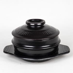 Può includere: Una pentola da cucina in ceramica nera con coperchio e piatto abbinato. La pentola ha una forma arrotondata con un coperchio che ha una piccola maniglia sulla parte superiore. Il piatto ha manici su entrambi i lati. La pentola e il piatto sono in ceramica nera lucida.