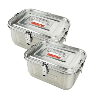 May include: Two stainless steel rectangular food storage containers with hinged lids and metal handles. The containers are stacked, with the top container slightly smaller than the bottom one. Each lid has a small label. The containers are made of shiny, brushed metal.