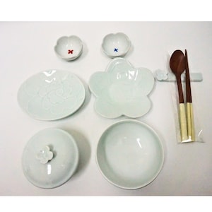 Puede incluir: Un conjunto de vajilla de cerámica verde pálido, que incluye platos, cuencos y un plato con tapa, dispuestos sobre una superficie blanca. El juego presenta un diseño floral e incluye palillos y cucharas de madera. Los cuencos pequeños tienen detalles rojos y azules.