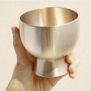 Könnte beinhalten: Eine messingfarbene Schale mit rundem Boden und schrägem Rand. Die Schale wird in einer Hand gehalten und zeigt ihre glatte, metallische Oberfläche. Das Design ist schlicht und elegant, geeignet zum Servieren oder Ausstellen.