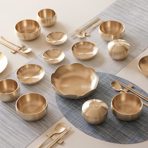 Peut inclure: Un assortiment de bols de service, de petites assiettes et de récipients à couvercle de couleur dorée. Des baguettes et des cuillères sont également incluses. Les articles sont disposés sur un set de table à motifs bleus et blancs et une surface claire.