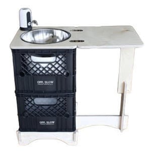 May include: A portable outdoor sink with a stainless steel basin and a white faucet. The sink is mounted on two black plastic crates and has a light-colored wooden countertop. The words "OFF. SLOW" are printed on the crates.
