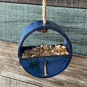 May include: A hanging bird feeder with a deep blue ceramic bowl and a natural rope hanger. The feeder is filled with birdseed and is mounted on a black metal bracket against a weathered wood background.