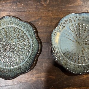May include: Two ceramic plates with a scalloped edge and a floral design. The plates have a blue and green glaze with a dark brown rim. The plates are on a dark wood surface.