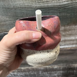 May include: A ceramic mug with a textured, uneven surface featuring a two-tone glaze. The upper portion is a mottled reddish-purple, while the lower section is a speckled cream colour. A white straw is inserted into a hole on the upper rim.