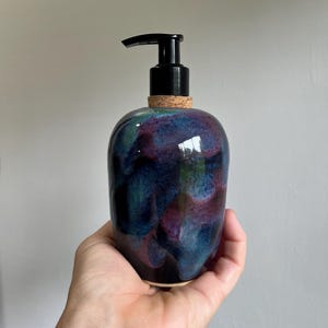 May include: A ceramic soap dispenser with a glossy finish. The dispenser features a black pump and a cork base. The body of the dispenser is a blend of dark blue, purple, and teal colors.