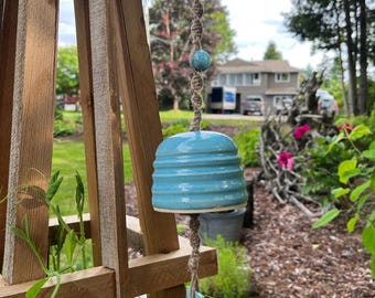 May include: A turquoise ceramic wind chime with a ridged bell shape hangs from a wooden trellis. The chime is suspended by a twisted rope, with a small turquoise bead and a flat, turquoise charm. The background features a garden setting.