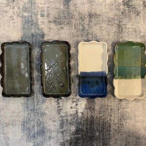 Op de afbeelding: Vier rechthoekige keramische trays met geschulpte randen. Elke tray heeft een uniek kleurenschema: een golvend patroon in grijsgroen, een getextureerd grijsgroen, wit en blauw en wit en groen. De trays zijn gerangschikt op een grijs en blauw patroon.